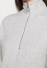 Gros plan sur un pull gris en maille chinée avec une fermeture éclair argentée à quart de tour et un anneau rond sur une personne aux cheveux foncés.