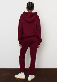 Rudo kapučino megztinis ir joggingo kelnės, minkšta medžiaga, laisvas siluetas, su gobtuvu, elastiniai rankogaliai ir juosmuo, derinami su baltais sportbačiais ir klasikinėmis kojinėmis.