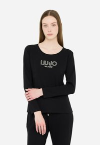 Noor noor sirged pruunid juuksed, seljas must pikakäiselise särgi, millel on "LIU�•JO MILANO" logo, seisab käed ristatud.