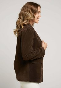 Blazer en daim marron avec un design structuré, fermeture à un bouton et deux poches avant. Texture lisse et coupe ajustée.