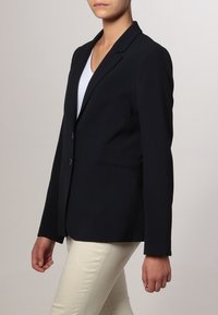 Blazer noir sur mesure avec revers crantés, deux boutons et poches. Porté sur une chemise blanche et associé à un pantalon clair. Texture lisse.