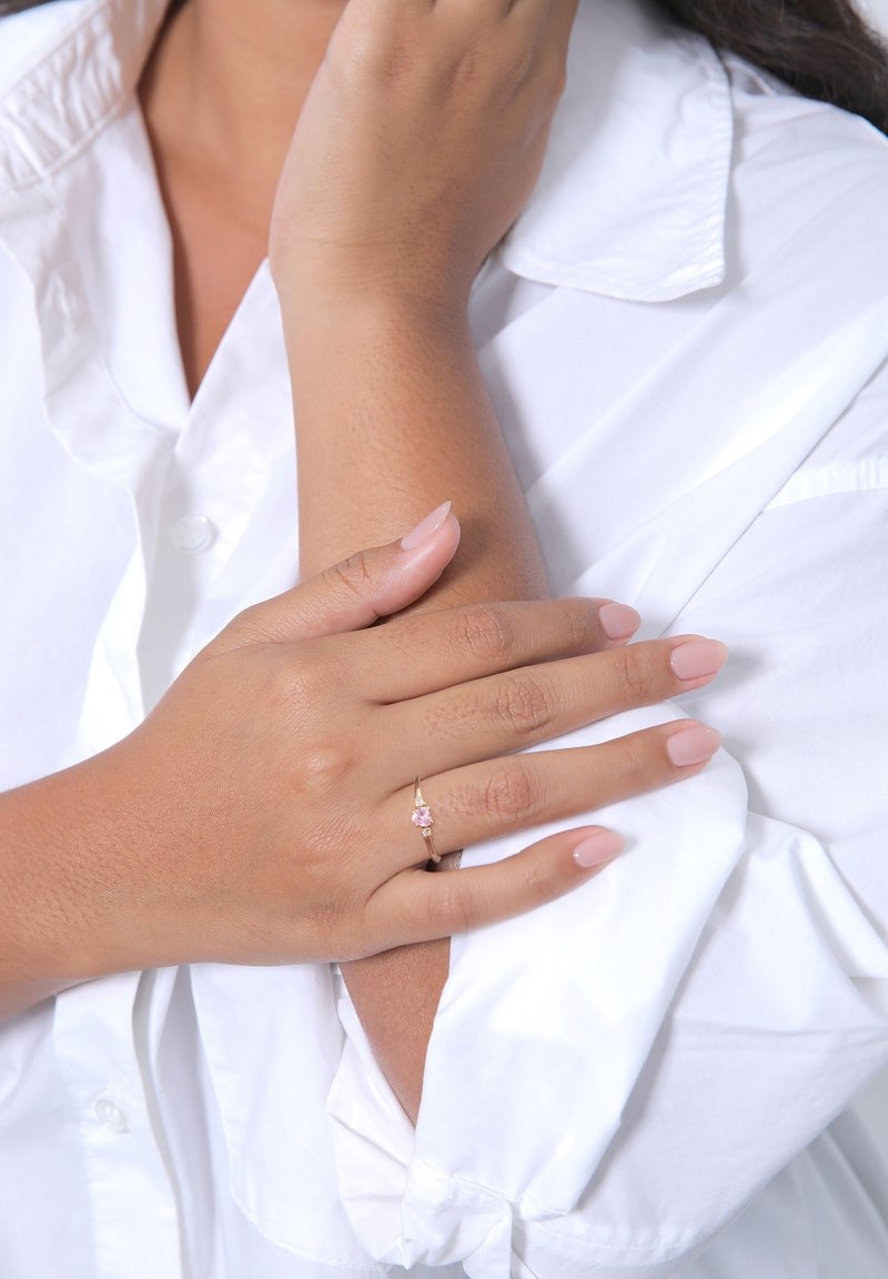 Femme portant une chemise blanche touche son bras opposé, montrant une bague en or avec une petite pierre précieuse rose sur l'annulaire de la main gauche.