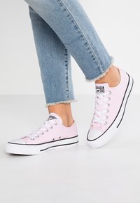 Osoba nosząca jasnoróżowe trampki Converse niskie oraz skończone jasnoniebieskie dżinsy z postrzępionymi nogawkami, stojąca na białej podłodze.