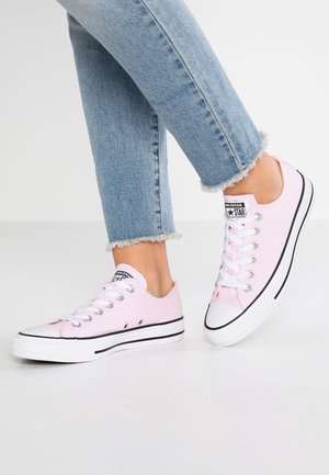 Person, die hellrosa Converse Low-Top-Sneaker und abgeschnittene hellblaue, fransige Jeans trägt, steht auf einem weißen Boden.