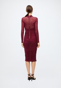 Robe moulante bordeaux avec manches longues transparentes, texture froncée, fermeture éclair au dos, et longueur au genou. Talons hauts noirs à lanières visibles.