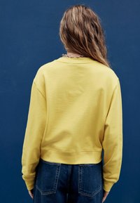 Geel sweatshirt met een ronde hals, losse pasvorm en geribbelde zoom, gecombineerd met donker denim jeans. De stof lijkt zacht met een lichte textuur.