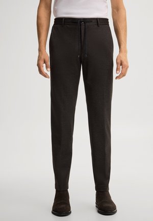 Mann trägt dunkelbraune, eng anliegende Hose mit Kordelzug in der Taille und dunkelbraune Wildlederschuhe vor schlichtem Hintergrund.