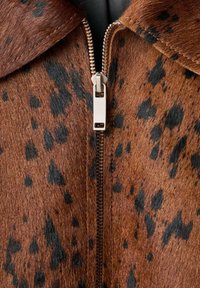 Veste en cuir marron avec un motif à pois, dotée d'une fermeture éclair en argent et d'un col. La texture semble douce et semblable à de la fourrure.