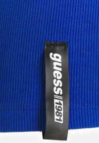 Blå ribbad tyg med en svart etikett som visar märkesnamnet "guess" och "1981" i vitt tryck, sytt vid kanten.