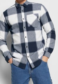 Camisa abotonada con patrón de cuadros en azul marino y blanco, mangas largas, un bolsillo en el pecho izquierdo y botones oscuros.