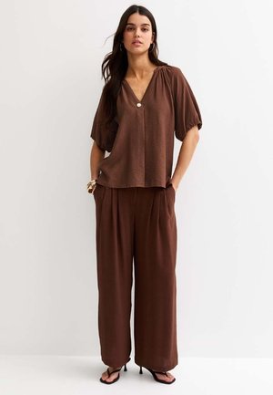 Kobieta nosząca luźną brązową bluzkę z dekoltem w serek i krótkimi bufiastymi rękawami, dopasowane szerokie spodnie, czarne sandały na obcasie oraz złote bransoletki.