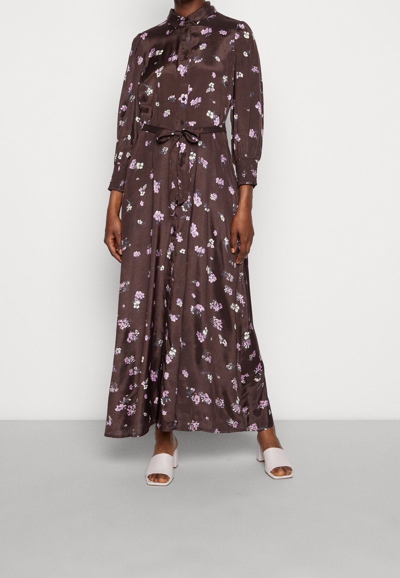 Braunblumiges Maxikleid mit langen Ärmeln, Knopfleiste, tailliertem Gürtel und weichem Satinmaterial. Getragen mit hellrosa offenen Schuhen.