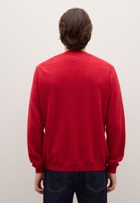 Roter Pullover aus glattem Strick mit langen Ärmeln, Rundhalsausschnitt sowie gerippten Bündchen und Saum, getragen zu dunkelblauen Jeans.
