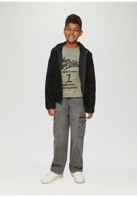 Zwarte fleece jack over een groene graphic t-shirt, grijze cargobroek met zijzakken en witte sneakers. Glimlachende jongentje staat tegen een lichte achtergrond.