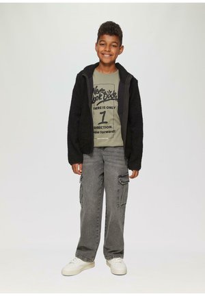 Zwarte fleece jack over een groene graphic t-shirt, grijze cargobroek met zijzakken en witte sneakers. Glimlachende jongentje staat tegen een lichte achtergrond.