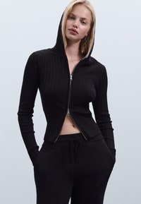 Crna rebrasta zip-up dukserica s prilagođenim dizajnom i kapuljačom. Sadrži srebrni zatvarač i duge rukave, u kombinaciji s odgovarajućim crnim trenirkama.