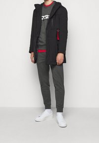 Parka negra con capucha, múltiples bolsillos, sudadera gris con acentos rojos, joggers grises y zapatillas blancas. Tejido texturizado en todo.
