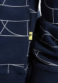 Tela de punto azul marino con patrones geométricos blancos, con una superficie texturada. Una pequeña etiqueta amarilla con un logo es visible en el lateral.