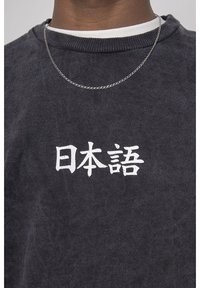 Osoba oblečená v černé mikině s bílými japonskými znaky a stříbrným náhrdelníkem přes bílém tričku s krátkým rukávem.