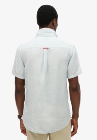 Hombre con camisa de manga corta azul clara con cuello levantado, de pie de espaldas, combinado con pantalones beige y un reloj de pulsera en el brazo izquierdo.