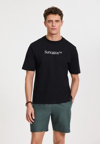 Fekete pamut póló rövid ujjal, fehér "Suncation™" logóval a mellkason, zöld sorttal párosítva.