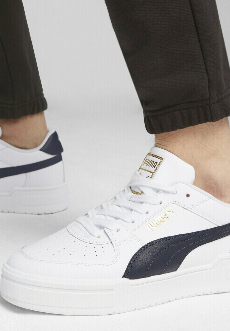 Personne portant des baskets blanches Puma avec une bande noire et un logo doré, associées à un pantalon noir à revers, debout sur une surface claire.