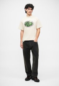 Beige cotton t-shirt with a green graphic design featuring an eye, paired with black loose-fitting jeans and dark shoes.