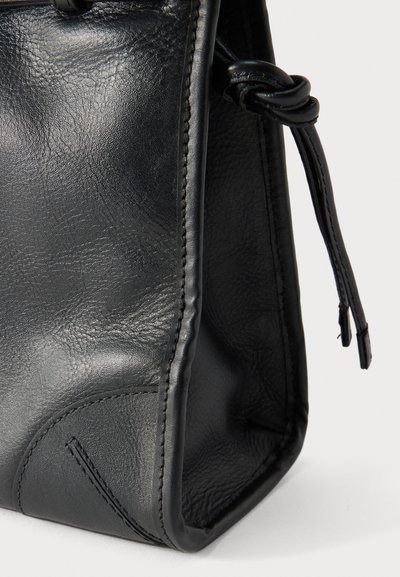 Coin d'un sac en cuir noir avec des coutures visibles et une lanière en cuir nouée sur un fond blanc.