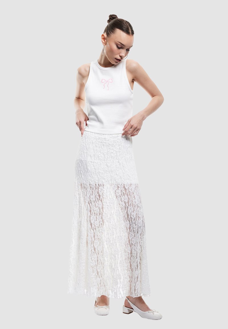 Witte geribbelde tanktop met roze strikborduursel, gecombineerd met een lange, doorzichtige witte kanten rok. Model draagt witte blokhak schoenen.