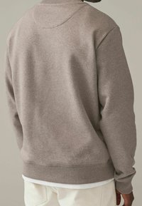 Hombre con sudadera marrón claro sobre camisa blanca y pantalones color crema, visto desde atrás contra un fondo liso.
