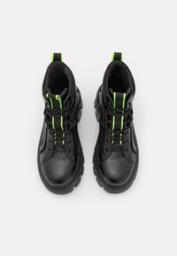 Bottes noires avec une semelle en caoutchouc, dotées de lacets et de coutures vertes. Le design comprend un col légèrement rembourré et des panneaux texturés.
