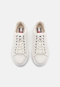Sneakers bianchi in tela con una superficie testurizzata, punta tonda, sei occhielli e suola in gomma. Presentano un'etichetta con accenti rossi, bianchi e blu.