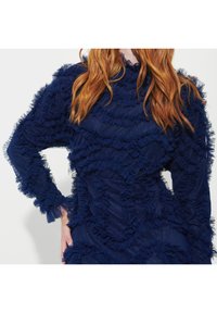 Marinblå klänning med volanglager och genomskinliga detaljer. Långa ärmar med frilldetaljer. Texturerad tyg skapar djup och visuell intresse.