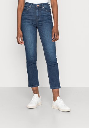 Personne portant un jean bleu taille haute coupé et des baskets blanches, debout devant un fond clair uni.