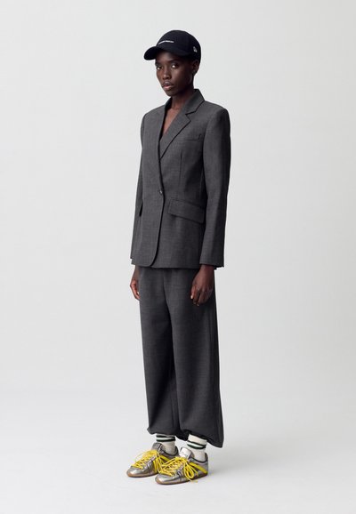 Traje gris a medida con chaqueta entallada, pantalones de pierna ancha, combinado con una gorra negra y zapatillas de plata con cordones amarillos y calcetines a rayas.