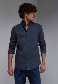 Camisa de botón azul marino hecha de tela suave, con un bolsillo en el pecho, mangas enrolladas y un cuello clásico. Combinada con jeans de mezclilla.