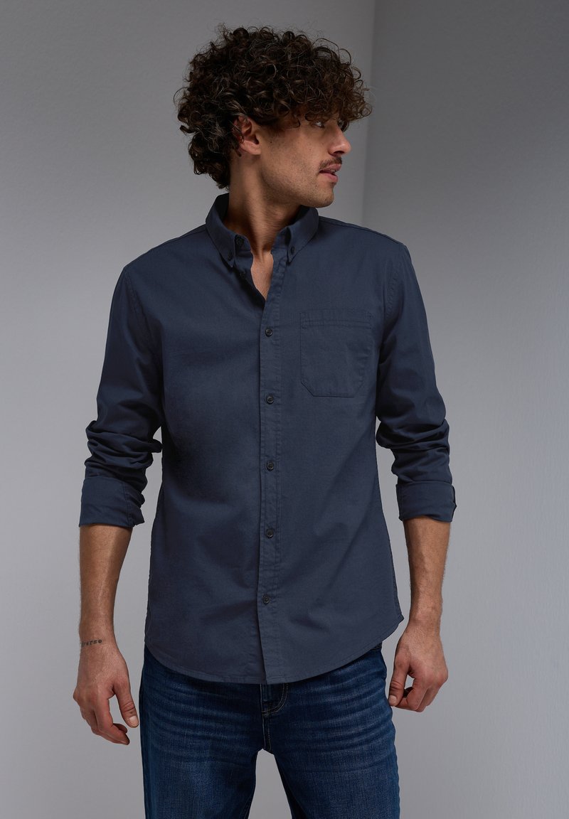 Camisa de botón azul marino hecha de tela suave, con un bolsillo en el pecho, mangas enrolladas y un cuello clásico. Combinada con jeans de mezclilla.