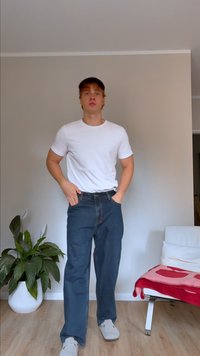 Camiseta blanca ligera, vaqueros oscuros de corte recto y zapatos gris claro. El fondo presenta una planta en maceta y una silla.