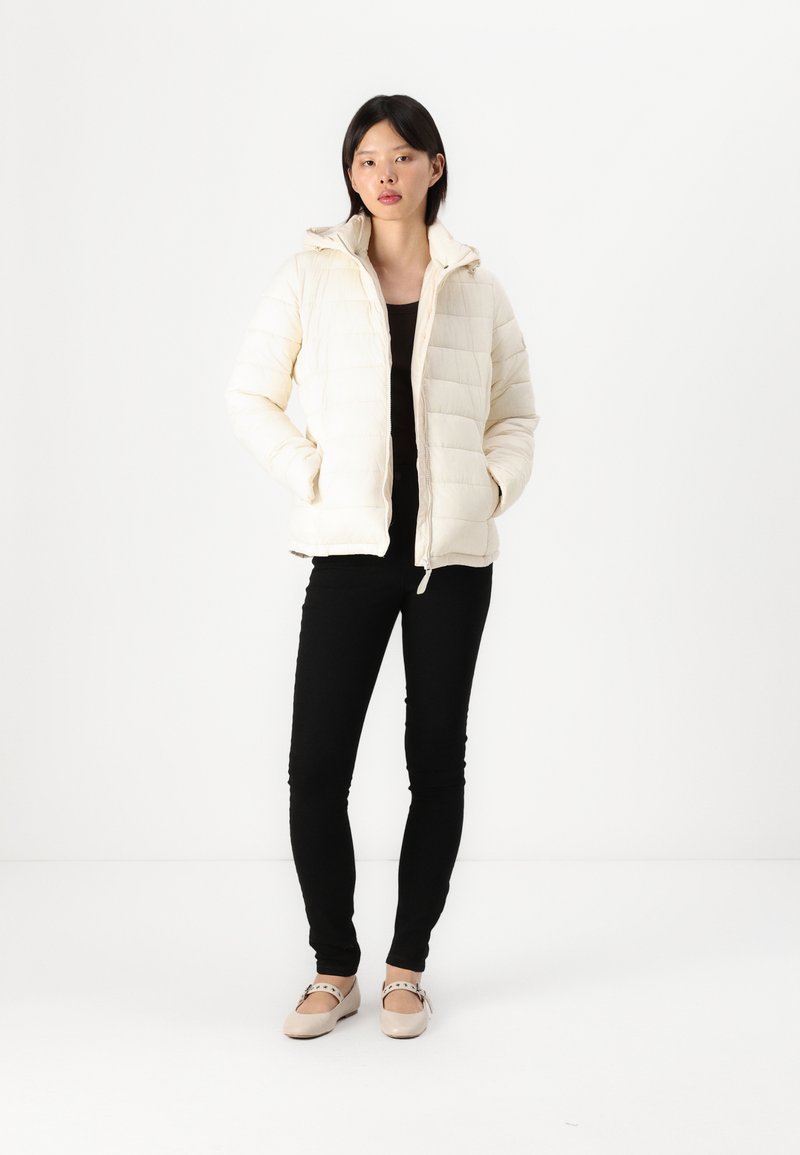 Giacca invernale di colore crema chiaro con texture trapuntata, zip frontale e cappuccio. Indossata sopra una maglia nera con pantaloni neri e scarpe chiare.