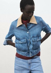 Denim jacket with beige collar, two front pockets, and snap buttons. Worn over a red shirt, paired with matching light blue jeans.