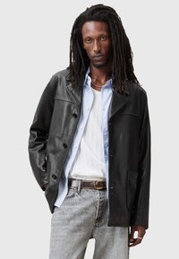 Schwarze Lederjacke mit Knopfleiste, einer Brusttasche und spitzen Revers, über einem hellblauen Hemd mit vertikalen Streifen getragen.