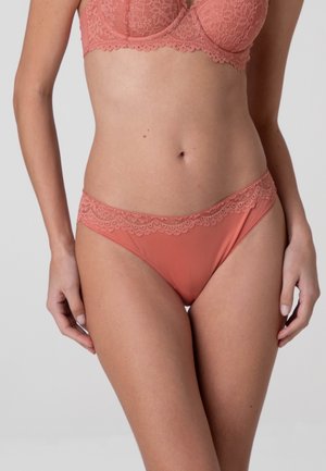 Donna che indossa un reggiseno in pizzo rosa corallo e slip abbinati con bordi in pizzo su uno sfondo chiaro e uniforme.