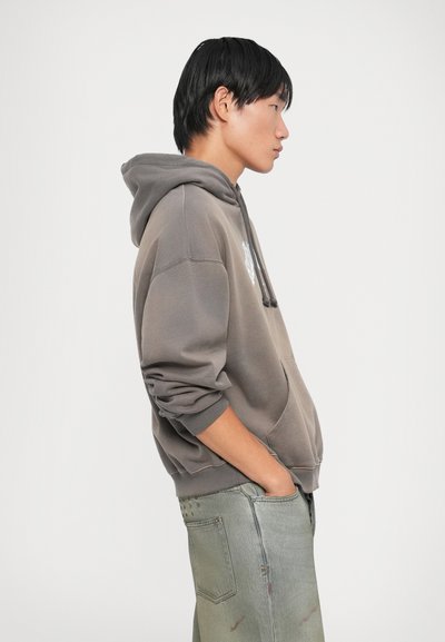 Sweat-shirt marron avec une poche avant et un cordon de serrage, porté sur un jean vert clair. Le sweat-shirt présente des poignets côtelés et une coupe décontractée.