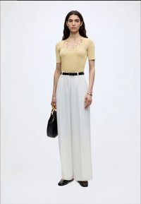 Γυναίκα με μακριά σκουρόχρωμα μαλλιά φοράει μπεζ κοντομάνικη μπλούζα με κέντημα, άσπρο παντελόνι καμπάνα με υψηλή μέση, μαύρη ζώνη, μαύρα παπούτσια, κρατάει μαύρη τσάντα.