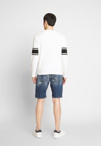 Vit ribbad tröja med svarta ränder på ärmarna, kombinerad med blå denimshorts. Modellen har på sig vita sneakers med marinblå detaljer.