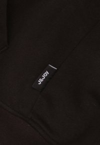 Matière de sweat noir avec une texture douce. Présente une petite étiquette avec "J&JOY" en blanc sur un fond noir. Design simple, sans motifs.