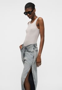 Mujer con gafas de sol negras, top sin mangas beige, y falda vaquera gris descolorida de tiro alto con abertura frontal, manos en los bolsillos, fondo neutro.