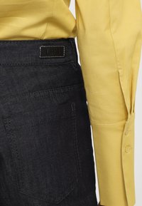 Mørk denim jeans med baklommer, med et svart merket på linningen. Overlappet av en tettsittende gul langermet skjorte.