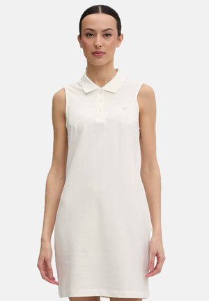 Donna che indossa un abito polo bianco senza maniche, in piedi davanti a uno sfondo bianco neutro, rivolta in avanti con espressione neutra.