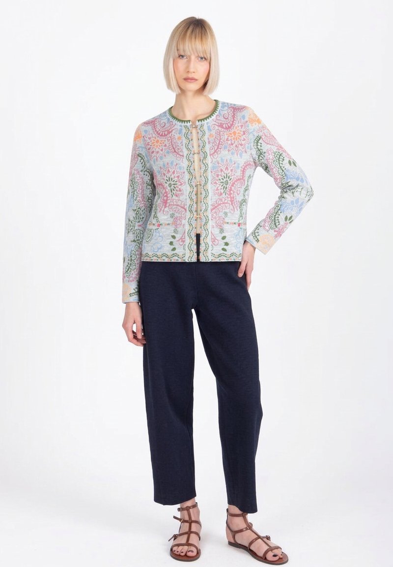 Lühikeste blondide juustega naine, kandes pastellsete mitmevärviliste mustritega jopet, tumesiniseid pükse ja pruune rihmikuid, seistes valge tausta ees.